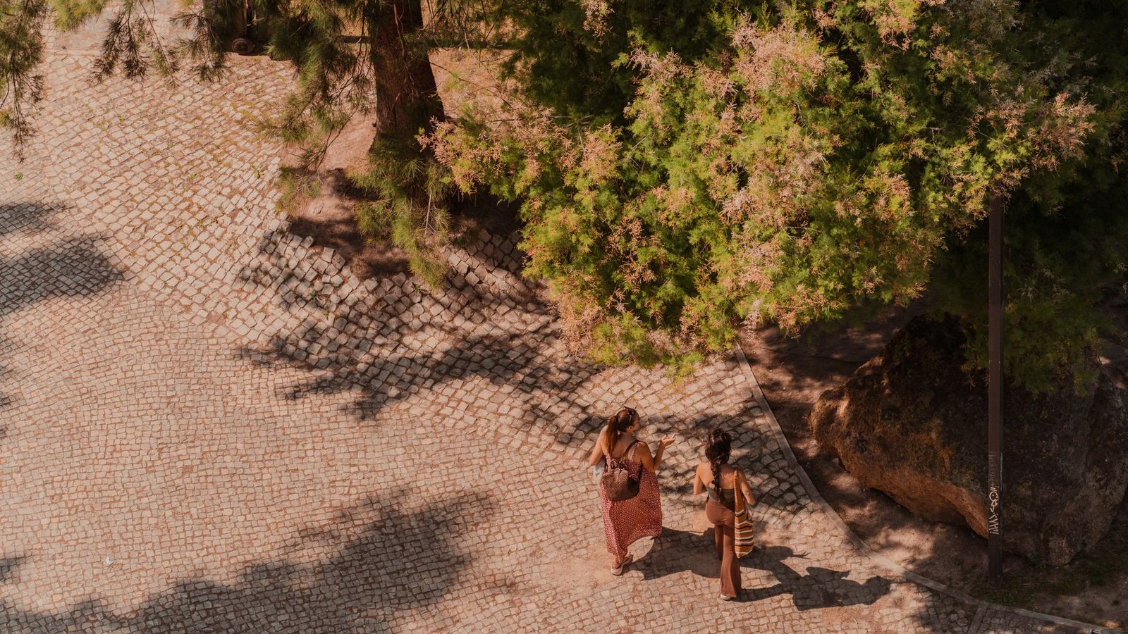 Aerial shot of two people walking on a cobblestone path in a sunny Lisbon park under trees.