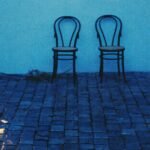 Two open doorways glowing warm amber at blue hour, their light reflected on wet cobblestones, with two empty bentwood chairs between them.