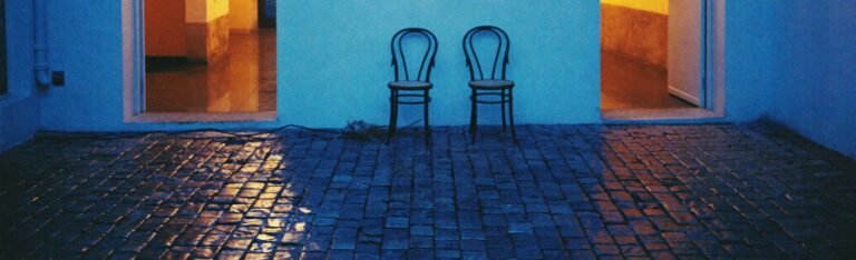 Two open doorways glowing warm amber at blue hour, their light reflected on wet cobblestones, with two empty bentwood chairs between them.