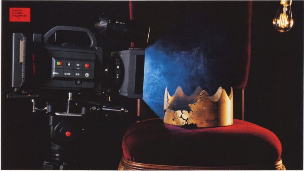 Still life of a broadcast camera on a tripod facing a cracked golden crown on a velvet theatre chair, lit by cold blue screen light against a dark backdrop — illustrating the collision of surveillance technology and theatrical power in SillySeason's Macbeth adaptation.