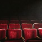 Rows of worn crimson velvet theatre seats in an empty auditorium, lit by a single raking shaft of stage light with dust suspended in the beam.