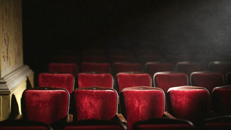 Rows of worn crimson velvet theatre seats in an empty auditorium, lit by a single raking shaft of stage light with dust suspended in the beam.